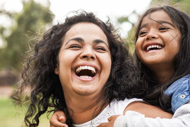 Two women smiling outdoors in a park, embracing joyfully together.