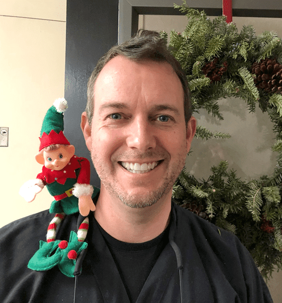 Man smiling at camera holding colorful toy elf, holiday decorations in background.