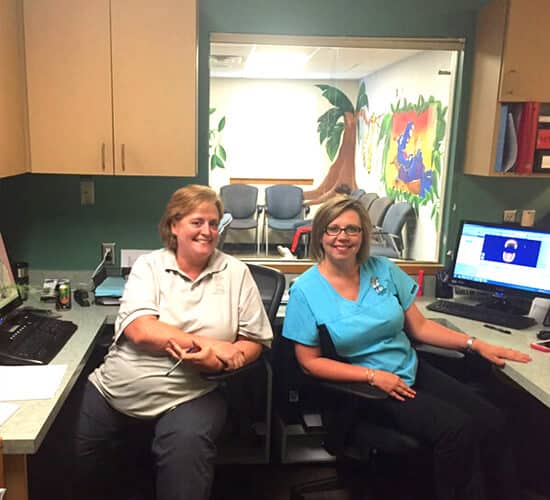 Two women sit at dental office desk with computers and equipment in background.