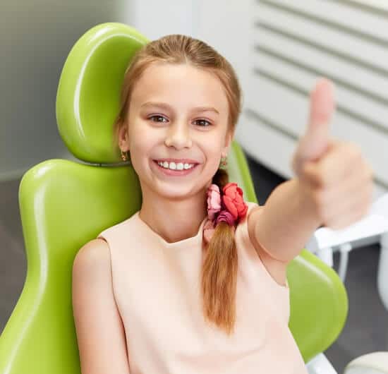 Young girl in dental chair giving thumbs up, smiling at orthodontist appointment.
