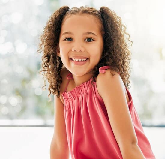 Young girl with curly hair smiling at camera, wearing pink top, bright indoor setting.