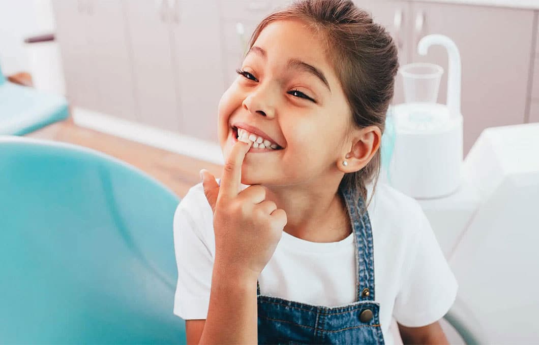 Young girl smiling in dental chair, touching her tooth during examination at dentist office.