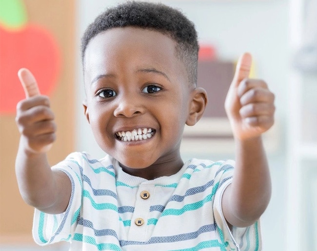 Smiling toddler in striped shirt giving double thumbs up in bright indoor setting.