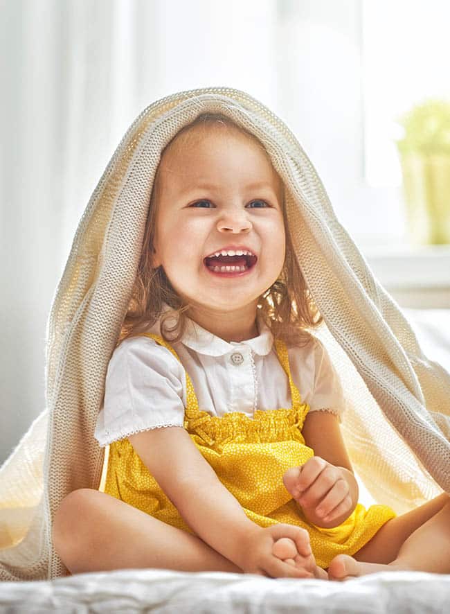 Happy toddler girl laughing under towel after bath in bright, sunny bathroom setting.