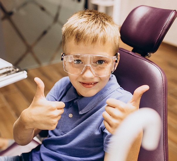 Young boy wearing glasses gives thumbs up in dental office chair, smiling.
