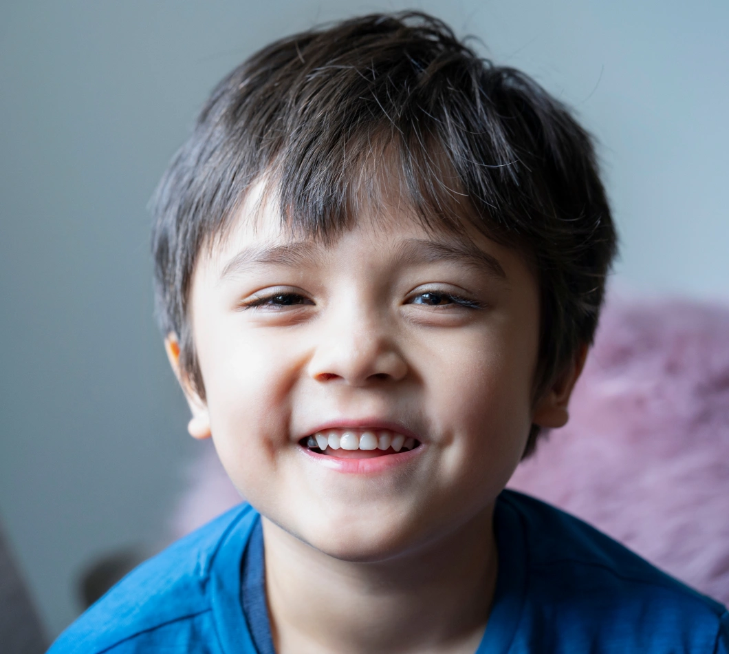 Young boy smiling brightly at camera wearing blue shirt against neutral background.